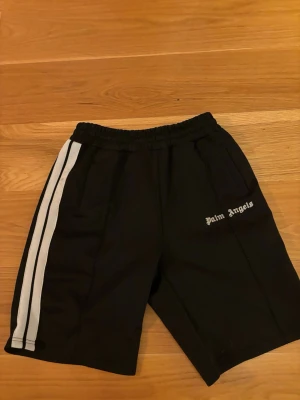 Svarta Palm Angels shorts med ränder - Svarta shorts från Palm Angels med vita dubbla sidoremsor och elastisk midja. Klassisk sportig look med fickor på sidorna och Palm Angels-logga broderad på vänster ben. Perfekta för en avslappnad streetwear-stil.