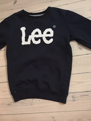 Mörkblå sweatshirt från Lee - Mörkblå sweatshirt från Lee med stort vitt logotryck på bröstet. Långa ärmar. Tröjan har ribbade muddar vid ärmslut och nederkant. Sparsamt använd. Storlek 10-11 år