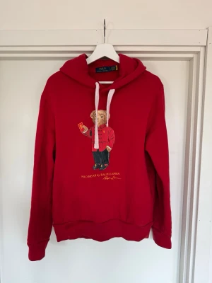 Röd hoodie Polo Bear Ralph Lauren - Röd hoodie från Ralph Lauren med Polo Bear-motiv på bröstet. Vita snören sitter i, men orginalsnöreb medföljer, en perfekt tröja för dig som vill ha nått att chilla i hemma eller gå för att gå ut att äta