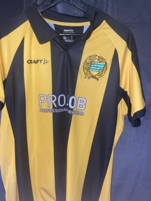 Hammarby tröja  - Snygg fotbollströja från Craft i gult och svart med vertikala ränder. Tröjan har klubbmärke med HIF på bröstet och tryck från Projob. Klassisk krage och korta ärmar, perfekt för match eller träning.