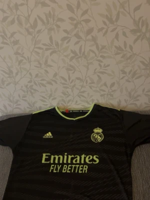 Real Madrid svart matchtröja Adidas - Snygg svart matchtröja från Real Madrid med neongula detaljer och klubbmärke på bröstet. Adidas-logga och texten 'Emirates Fly Better' på framsidan. Perfekt för fotbollsträning eller att bära till vardags.