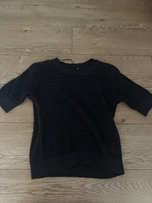 Marin blå stickad tröja med kort ärm - Aldrig använd!! Mörk blå stickad tröja med korta ärmar och rund halsringning. Tröjan har ett luftigt, hålmönstrat stickat material och breda muddar vid ärmslut och nederkant. Perfekt för lager på lager och enkel att matcha till olika outfits.