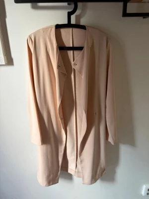 Ljusbeige lång kappa med tryckknappar - Stilren ljusbeige kappa i ett tunt, mjukt tyg. Modellen är rak och lång med tryckknappar framtill och utan synliga fickor. Perfekt för lager på lager och enkel att matcha med olika outfits. Passar dig som gillar minimalistisk och clean stil.