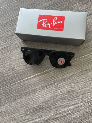 Svarta Ray-Ban Wayfarer solglasögon - Snygga svarta Ray-Ban Wayfarer solglasögon med polariserade linser. Klassisk fyrkantig form och diskret logga på glaset. Perfekta för att ge din stil en cool touch och skydda ögonen mot solen.