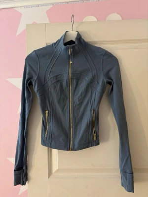 Blå träningsjacka från Lululemon - Slimmad blå träningsjacka från Lululemon med guldiga dragkedjor och hög krage. Jackan har två fickor med dragkedja och tumhål vid ärmsluten. Perfekt för gymmet eller löprundan, tillverkad i stretchigt funktionsmaterial.