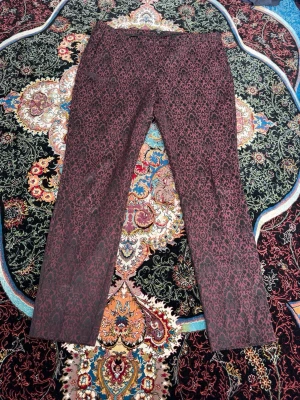 Vinröda kostymbyxor med mönster - Säljer ett par vinröda kostymbyxor med svart barockinspirerat mönster. Byxorna har rak passform och sidfickor. Materialet känns mjukt och har en elegant struktur som sticker ut. Perfekta för dig som vill ha något unikt och stilrent.