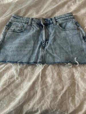 Blå jeanskjol med fransig kant - Säljer en kort blå jeanskjol med rå, fransig kant och klassiska fyra fickor. Kjolen har bälteshällor och dragkedja framtill. Perfekt för dig som gillar en avslappnad och trendig stil.