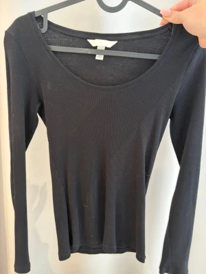 Svart ribbad långärmad topp - En svart, ribbad långärmad topp med rundad urringning. Toppen har en slim passform och är tillverkad i ett mjukt bomullsmaterial som känns skönt mot huden. Perfekt att styla med jeans eller kjol för en clean look.