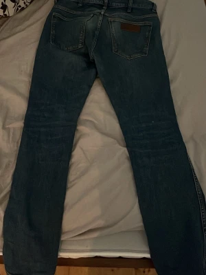 Wrangler jeans - Säljer ett par mörkblå jeans med rak passform och klassisk femficksdesign. Jeansen har bälteshällor och en läderpatch bak i midjan. Materialet är denim i bomull och de har en snygg tvättad look.