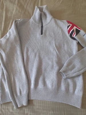 Grå halvzip tröja Henri Lloyd - Snygg grå stickad tröja från Henri Lloyd med halvzip och hög krage. På ena ärmen finns en stor Union Jack-detalj i rött, vitt och blått samt broderad logga. Perfekt för dig som gillar klassisk brittisk stil och vill ha något bekvämt och stilrent nu när kvällarna börjar bli kallare. 