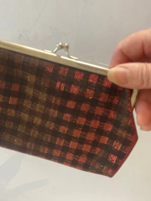 Rutig kuvertväska  - Snygg kuvertväska med rutigt mönster i rött, guld och svart. Väskan har metallram och klassiskt trycklås. Materialet skimrar lätt och ger en cool vintagekänsla. Perfekt accessoar för att lyfta din outfit.