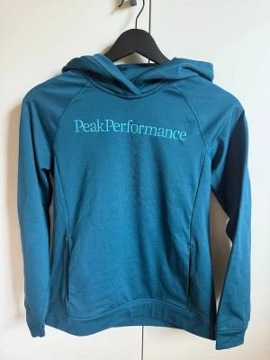 Blå hoodie från Peak Performance - Snygg blå hoodie från Peak Performance med logga broderad på bröstet. Hoodien har huva, långa ärmar och en magficka. Tillverkad i mjukt och stretchigt material som känns skönt mot huden. Perfekt för en avslappnad och sportig stil. Bara använd några enstaka gånger.