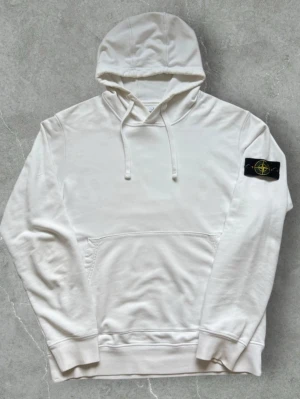 Stone Island Hoodie Vit - Snygg vit hoodie med huva och snörning samt en stor magficka. På vänster ärm sitter en svart patch med gult och grönt broderat märke. Hoodien har ribbade muddar vid ärmslut och nederkant, perfekt för en clean och stilren look.