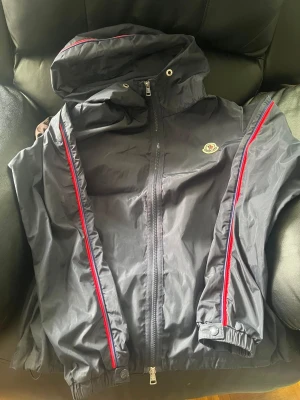 Moncler hattab vindjacka med huva - Säljer min jacka eftersom jag inte har något behov av den. Den har dessutom scan på Moncler loggan som tar dig direkt till monclers hemsida. Marinblå vindjacka från moncler med huva och dragkedja framtill. Jackan har röda och blå ränder längs ärmarna och ett Moncler-märke på bröstet. Tillverkad i lätt, glansigt material och har elastiska muddar vid ärmslut och nederkant.