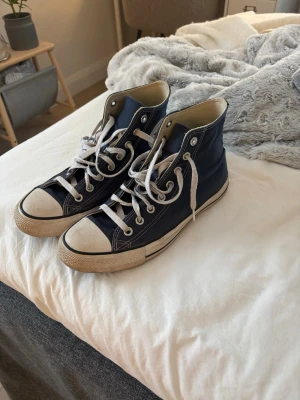 Converse All Star High - Klassiska mörkblå Converse All Star high-top sneakers med vita snören. Ovandelen är i canvas och sulan är vit med svart rand. Perfekta för en avslappnad och cool stil. Använd fåtal gånger.