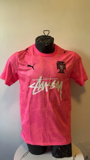 Rosa Stüssy x Puma fotbollströja - Säljer en rosa fotbollströja från Puma i samarbete med Stüssy. Tröjan har korta ärmar, svart Puma-logga på bröstet och axlarna, samt Stüssy-logga i vitt framtill. Detaljerat mönster i tyget och klubbmärke på vänster bröst.
