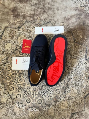 Marinblå sneakers från Christian Louboutin - Snygga marinblå sneakers från Christian Louboutin med klassisk röd sula och rund tå. Skorna har ovandel i mocka och textil, samt matchande marinblå skosnören. Perfekta för dig som vill sticka ut med exklusiv design och ikoniska detaljer.
