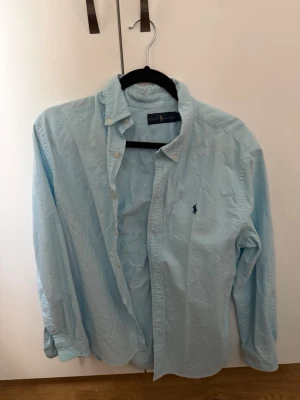 Ljusblå skjorta från Ralph Lauren - Klassisk ljusblå skjorta från Ralph Lauren med button-down krage och broderad logga på bröstet. Skjortan har långa ärmar och knappar framtill. Perfekt till jeans eller chinos för en clean och stilren look.