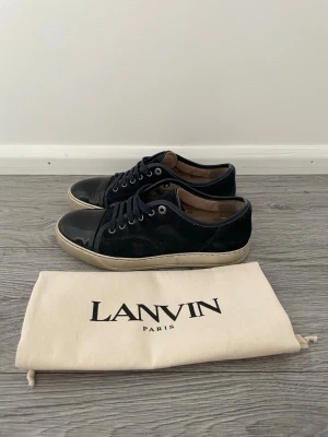 Mörkblåa lanvins med glans. UK=6 passar 41 - Snygga sneakers från Lanvin med ovandel i mocka och lackad tå. Skorna har vita sulor, mörkblåa skosnören och metallfärgade öljetter. Klassisk låg modell som passar till många olika stilar. Medföljer originalpåse och box.