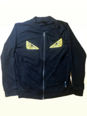 Svart Fendi bug eyes tröja - Cool svart hoodie med dragkedja och två stora gula ögonmotiv med svarta detaljer på bröstet. Hoodien har långa ärmar och ribbade muddar. Själva bug eyes motivet designades utav Karl Lagerfeld och är en galen grail i mode världen. Tveka inte på att höra av er vid några frågor!