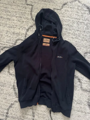 Svart hoodie med dragkedja och huva - Snygg svart hoodie från Parajumpers med dragkedja framtill och broderad 'PJS' logga på bröstet. Hoodien har justerbar huva med snören, ribbade muddar och fickor på sidorna. Insidan har en orange detalj vid nacken för extra stil. Använd ngra gånger men helt ny typ ngra små trådar som stycker ut men dom syns knapp