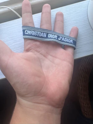 Christian Dior J'Adior blått armband - Säljer ett snyggt blått tygarmband från Christian Dior med texten 'J'Adior' broderad i vitt. Armbandet har dekorativa tofsar i ändarna och är perfekt för dig som gillar exklusiva accessoarer med streetstyle-vibe. VID FLERA ARMBAND KÖP KAN PRIS DISKUTERAS!