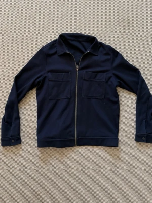 Mörkblå overshirt med dragkedja - Massimo dutti? - Stilren mörkblå overshirt med två stora bröstfickor och dragkedja framtill. Overshirten har klassisk krage och knappar vid ärmsluten. Materialet ser ut att vara mjukt och lite tjockare, perfekt för lager på lager. Glömt vad för märkte det är! Men köpte den på plick förra året för 700 kr och säljer den nu för 500 men pris är alltid diskuterbart! Tror märket var Massimo dutti
