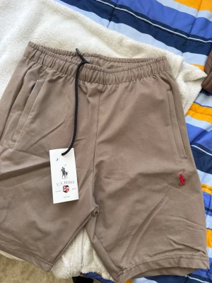 Beige shorts från U.S. Polo Assn - Snygga beige shorts från U.S. Polo Assn med elastisk midja och snörning. Shortsen har två sidofickor och en liten röd logga broderad på vänster ben. Perfekta för sommardagar och avslappnade tillfällen.