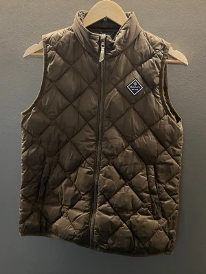 Brun quiltad dunväst från Gant - Snygg brun dunväst från Gant med quiltat mönster och hög krage. Västen har dragkedja framtill, två sidofickor och en diskret logotyp på bröstet. Perfekt för lager på lager och ger en sportig look.