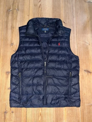 Mörkblå dunväst från Polo Ralph Lauren - Snygg mörkblå dunväst från Polo Ralph Lauren med hög krage och dragkedja framtill. Västen har två fickor med dragkedja och den klassiska röda loggan broderad på bröstet. Perfekt för lager på lager och riktigt stilren look. En mindre lagning vid magen men syns knappt då den lagats med samma färg som västen, utöver de helt nyskick!      Nypris:2195kr.   Mitt pris:800kr