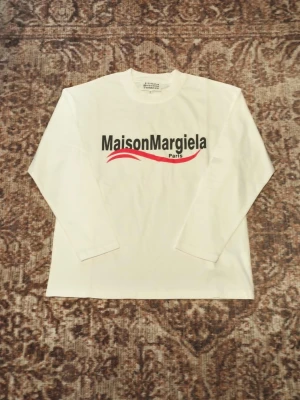 Vit långärmad topp Maison Margiela - Vit långärmad topp från Maison Margiela med tryckt logga i svart och rött framtill. Tillverkad i mjuk bomull och har en klassisk rund halsringning. Perfekt för dig som gillar minimalistisk och stilren design.