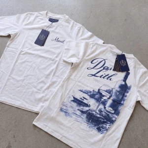 Vit t-shirt med blått tryck - Vit t-shirt med blått tryck på ryggen som föreställer en vinflaska, vinglas och båt vid vattnet. På bröstet står det 'Marcel' i blå text. T-shirten har rund hals och korta ärmar, perfekt för en avslappnad stil.