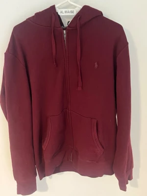 Vinröd hoodie med dragkedja från Polo Ralph Lauren - Säljer en vinröd hoodie från Polo Ralph Lauren med dragkedja framtill och klassisk broderad logga på bröstet. Hoodien har huva med dragsko, kängurufickor och ribbade muddar. Perfekt för en chill och stilren look.