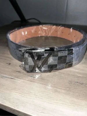 Rutigt bälte med metallspänne - Snyggt bälte med rutigt mönster i svart och grått samt ett stort metallspänne framtill. Insidan är beige och bältet har flera hål för justering. Perfekt accessoar för att lyfta din outfit.