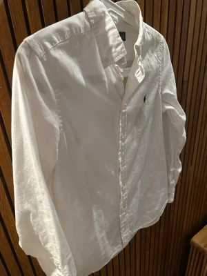 Vit skjorta från Ralph Lauren - Klassisk vit skjorta från Ralph Lauren med button-down krage och broderad logga på bröstet. Skjortan har långa ärmar och knäppning framtill. Perfekt till jeans eller chinos för en clean och tidlös look.