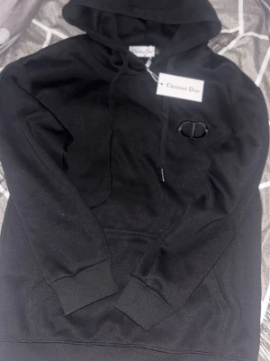 Zara - Snygg svart hoodie med huva och dragsnören. På bröstet finns en diskret broderad logga i svart. Hoodien har en klassisk känguruficka framtill och ribbade muddar vid ärmar och nederkant. Perfekt för en avslappnad och trendig stil.