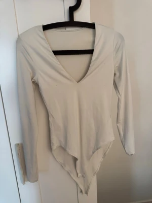 Vit långärmad body från Zara - Snygg vit body från Zara med lång ärm och djup V-ringning. Tillverkad i mjukt stretchigt material som sitter skönt mot kroppen. Perfekt att styla med jeans eller kjol för en clean look.
