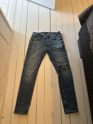 Jack & Jones - Snygga blå slim fit jeans från Jack & Jones med slitningar och hål på ena benet för en trendig look. Klassisk femficksmodell med dragkedja och knapp. Perfekta för dig som gillar en avslappnad men stilren stil.