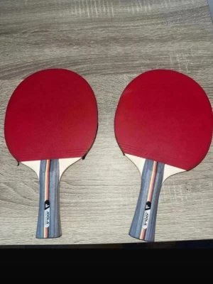 Två pingisracket från Joola - Säljer två klassiska pingisracket från Joola med röd och svart gummiyta. Handtagen har en snygg randig design i blått, rött och vitt. Perfekta för dig som vill spela bordtennis med stil och bra grepp.