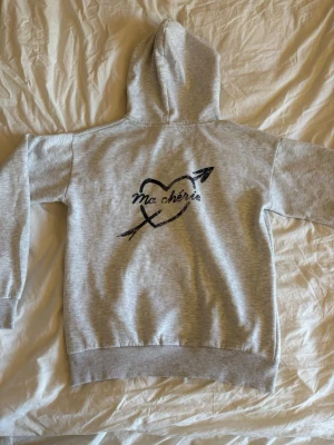 Fel kategori – ingen jeansprodukt - Säljer en ljusgrå hoodie med huva och trycket 'Ma chérie' i svart på både bröstet och ryggen. Ryggen har även ett hjärta med pil runt texten. Hoodien har ribbade muddar vid ärmar och nederkant. Perfekt för en chill och avslappnad stil. Storlek 158-164 men skulle klassa som XS