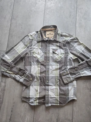 Grå och vit rutig skjorta Hollister - Säljer en grå och vit rutig skjorta från Hollister med klassisk krage. Skjortan har långa ärmar och detaljer i form av knappar framtill och på ärmsluten. Perfekt för en avslappnad och stilren look. Kom med prisförslag och frågor!