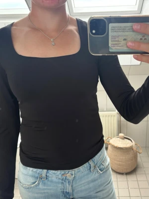 Svart långärmad topp med fyrkantig ringning - En stilren svart långärmad topp med fyrkantig ringning. Toppen har en tight passform och är tillverkad i ett mjukt, stretchigt material som sitter snyggt på kroppen. Perfekt att matcha med jeans eller kjol för en clean look.