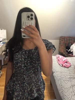 Blommig topp med volang och kort ärm - Söt blommig blus i mörkblått med små färgglada blommor. Modellen har volangkant nertill, smockad midja och korta ärmar med lätt rynk. Knappar framtill. Tunn och luftig, perfekt för sommaren.