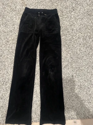 Svarta juicy couture mjukisar - Säljer ett par svarta mjukisbyxor i mjuk velour med raka ben och bred resår i midjan. Byxorna har snörning i midjan och stora fickor fram samt bakfickor med knappdetaljer. Perfekta för en chill och avslappnad stil.