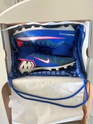 Blå Nike Air Zoom fotbollsskor storlek 43 - Säljer ett par blå Nike Air Zoom fotbollsskor med vita och rosa detaljer samt en hög krage för extra stöd. Skorna har en modern design med tydliga loggor och mönster på sidan. Perfekta för dig som vill sticka ut på planen och få bra grepp på konstgräs. Storlek 43. Sparsamt använda 