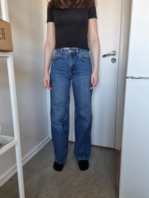 Blå raka jeans med hög midja - Säljer ett par klassiska blå jeans med rak passform och hög midja. Jeansen har fem fickor, bälteshällor och stängs med knapp och dragkedja. Materialet är kraftig denim i 100% bomull. Perfekta till en enkel t-shirt eller hoodie.