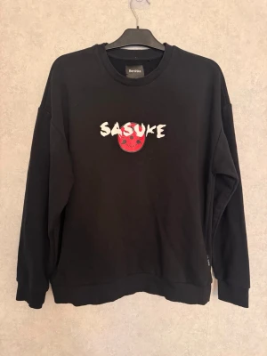 Svart Naruto Sasuke stickad tröja - Cool svart stickad tröja från Bershka med Naruto Shippuden-tema. På bröstet står det 'Sasuke' i vitt med en röd Uchiha-klan symbol. Tröjan har rund hals och långa ärmar, perfekt för dig som älskar anime och streetwear.