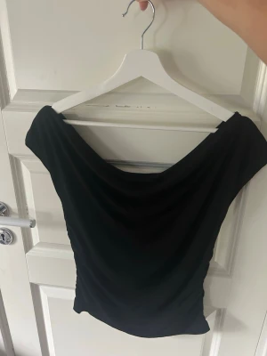 Svart offshoulder topp - Snygg svart offshoulder topp med draperad halsringning. Toppen är tight och har en stilren look som passar perfekt till jeans eller kjol. Tillverkad i ett mjukt och stretchigt material för en bekväm passform.