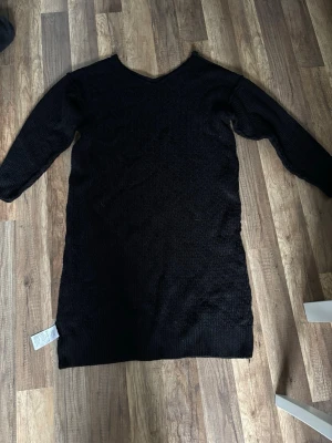 Svart stickad lång tröja - Mysig svart stickad lång tröja med V-ringning och långa ärmar. Tillverkad i 100% akryl vilket gör den mjuk och lätt att ta hand om. Perfekt att styla med tights eller jeans för en avslappnad look.