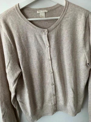 Beige kofta från H&M Basic - Klassisk beige kofta från H&M Basic med rund halsringning och knäppning framtill. Tillverkad i mjukt stickat material som känns skönt mot huden. Perfekt att slänga över en topp eller klänning för en enkel och clean look. Tar plats i garderoben bara.